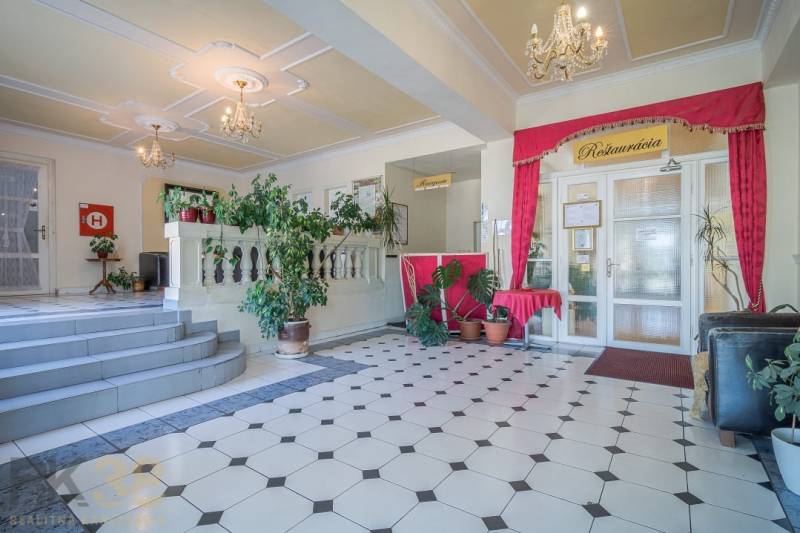 Hotelová recepce s rostlinami a lustry, dlažba s geometrickým vzorem.