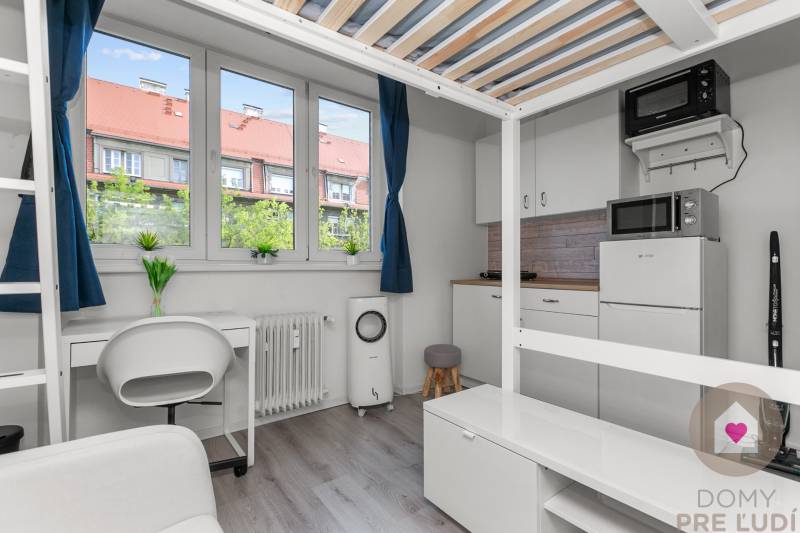 Garsonka s podlahou s dřevěným dekorem, pracovním stolem a kuchyní s mikrovlnnou troubou.