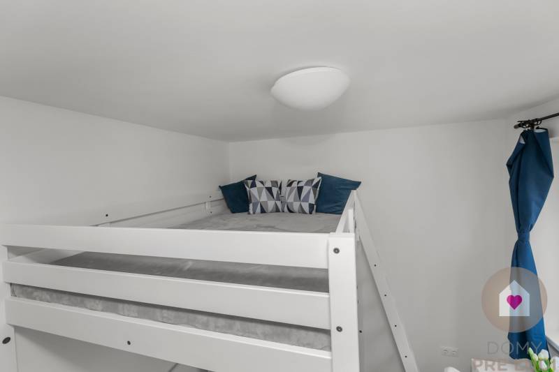 Postel na vyvýšeném místě v garsonce s polštáři a modrým závěsem.