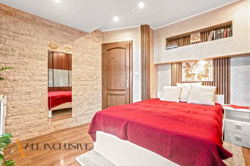 Ložnice v rodinném domě s postelí, kamennými detaily a podlahou s dřevěným dekorem.