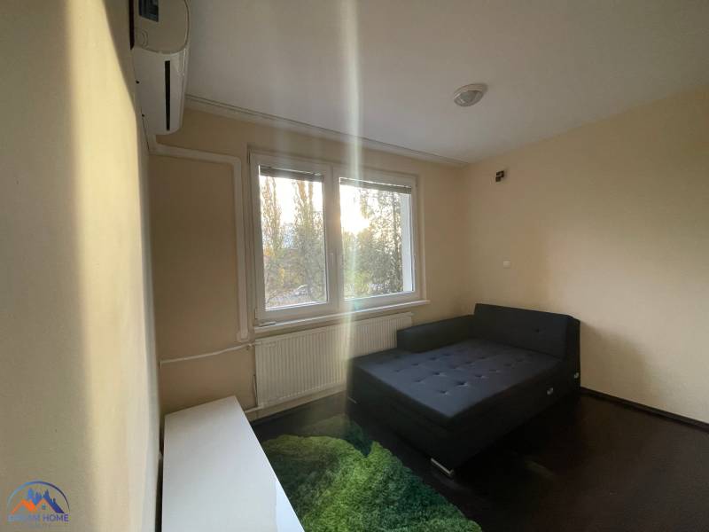 Jednoduchý pokoj s postelí, kobercem a oknem v jednopokojovém bytě.