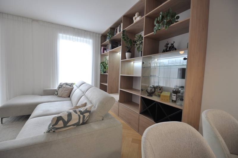 Obývací pokoj v rodinném domě s bílou pohovkou, dřevěnou knihovnou a rostlinami.