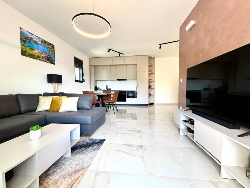 Obývací pokoj jednopokojového bytu s pohovkou, TV a kuchyňským koutem s posezením.