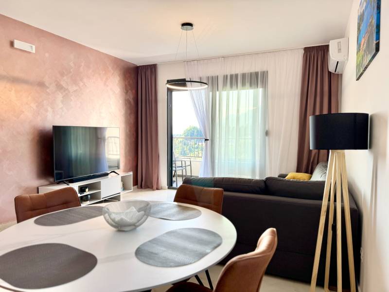 Obývací pokoj v jednopokojovém bytě s televizí, balkonem a elegantním stolem.