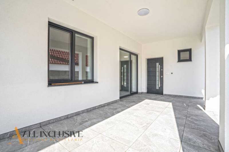 Rodinný dům s terasou, velkými okny a šedou podlahou s dlažbou.