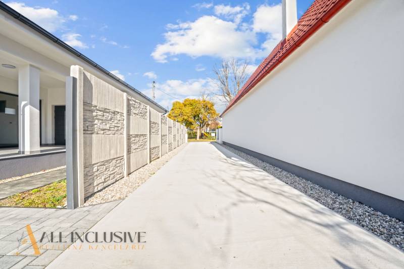 Úzká příjezdová cesta mezi rodinnými domy v Orechové Potôni, lemovaná zdí a stromy.