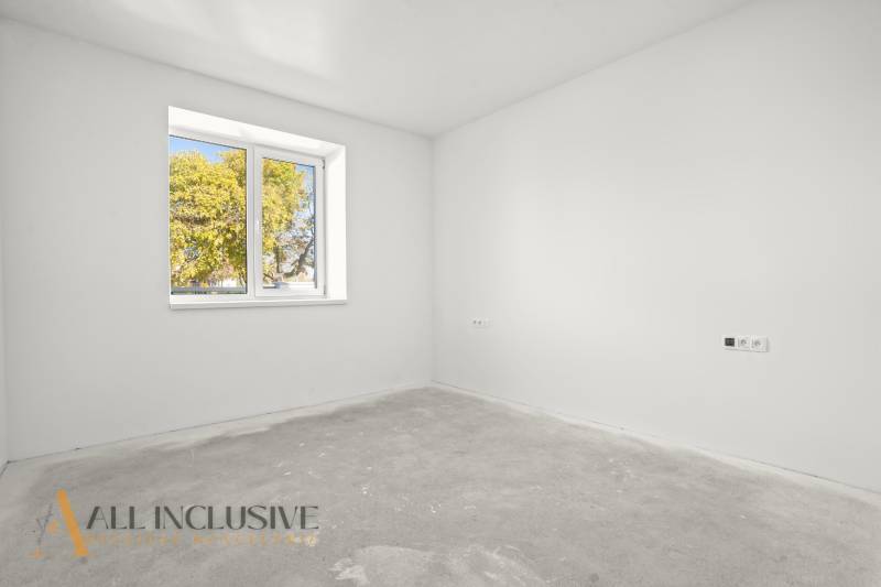 Prázdná místnost v rodinném domě s bílými stěnami a oknem s výhledem na stromy.