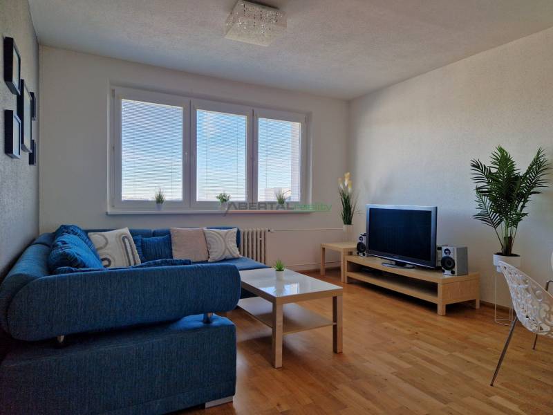 Obývací pokoj v 3-pokojovém bytě s modrou pohovkou, televizí a podlahou s dřevěným dekorem.