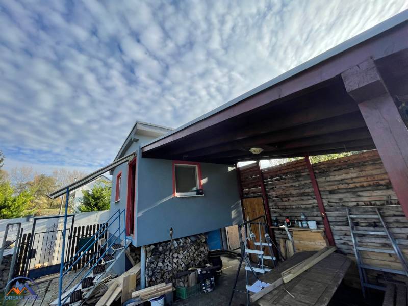 Chata v Komárně na Nové osadě se schodištěm a dřevěným přístřeškem pod oblaky.