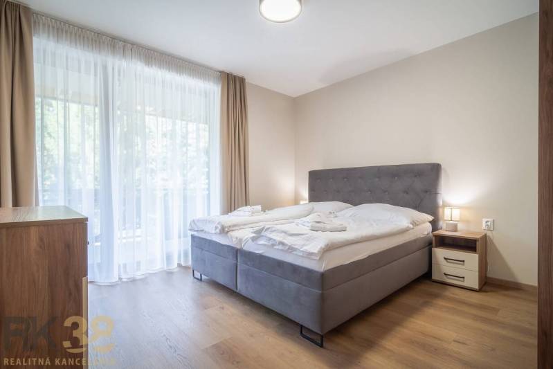 Ložnice v 2pokojovém bytě se světlými závěsy a podlahou s dřevěným dekorem.