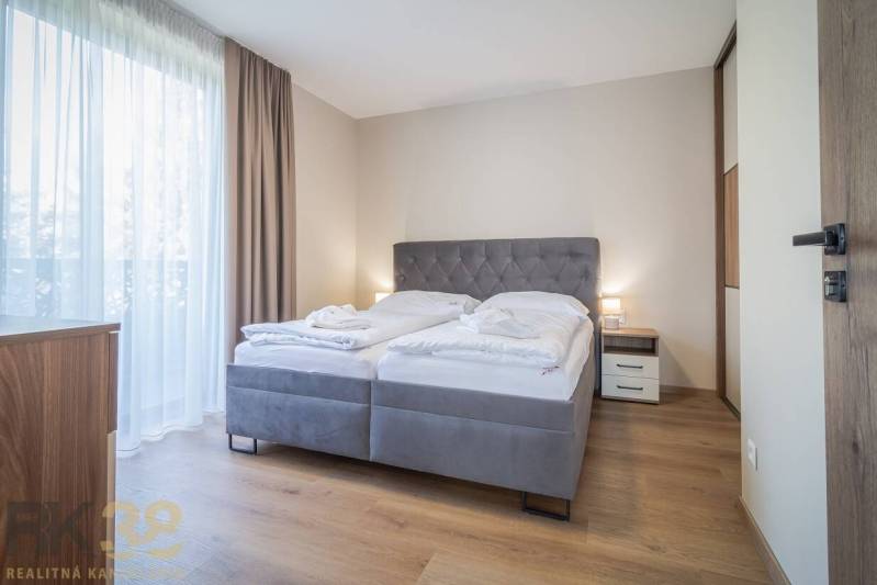 Ložnice v 2pokojovém bytě s pohodlnou postelí, podlahou s dřevěným dekorem a závěsy.