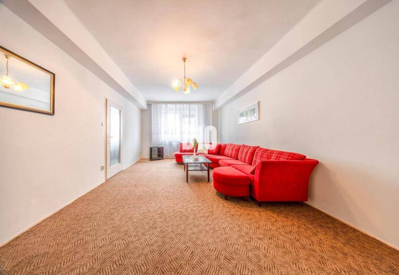 Obývací pokoj se světlou stěnou, červenou sedačkou a kobercem ve 4pokojovém bytě.