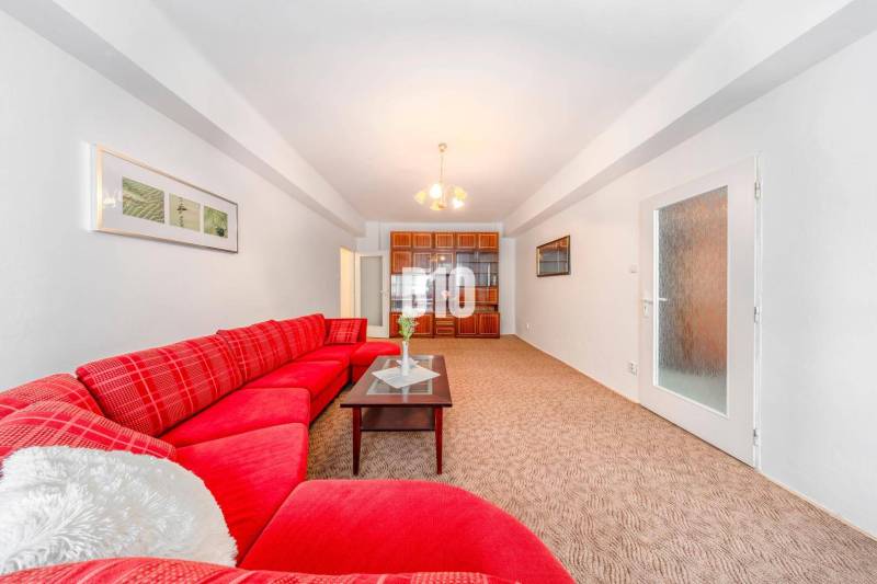 Červená pohovka, skleněný stolek, vestavěná skříň a světlý koberec ve 4pokojovém bytě.