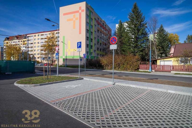 Exteriér s bytovkou a parkovištěm v Popradu, v pozadí zeleň a domy.