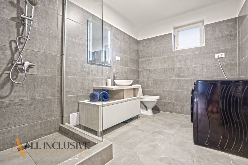 Koupelna v rodinném domě s dlaždicemi, sprchou, pračkou a umyvadlem.