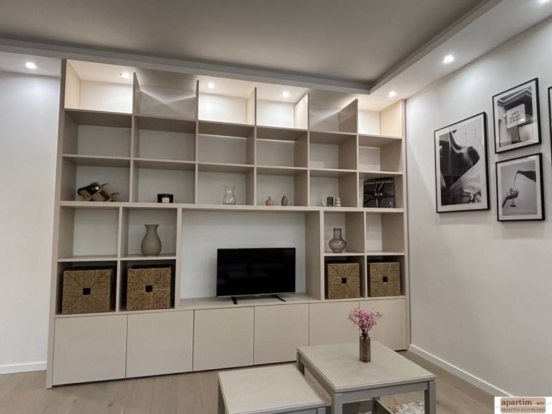 Úložná stěna s policemi a dekoracemi v 2-pokojovém bytě, vedle stůl s květinami.