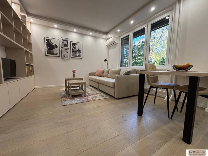 Obývací pokoj ve 2pokojovém bytě, velká sedačka, stolek, podlaha s dřevěným dekorem.