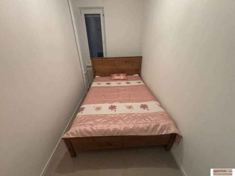 Úzká ložnice v 2-pokojovém bytě s dřevěnou postelí a květinovým přehozem.