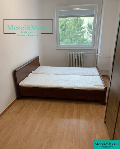 Ložnice s postelí, podlaha s dřevěným dekorem, okno s výhledem na zeleň.