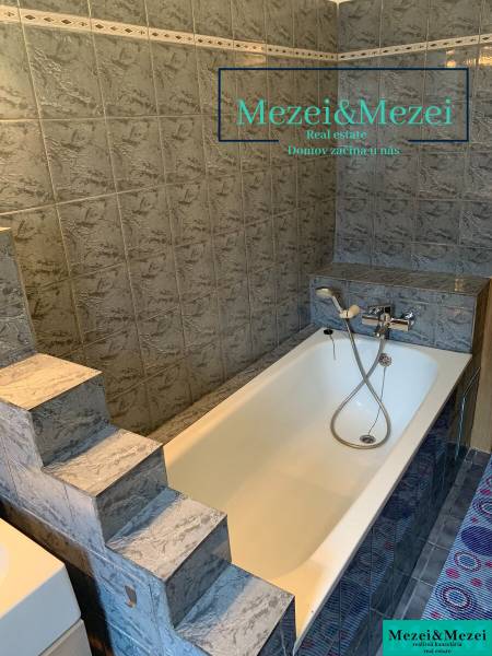 Koupelna se schůdky a vanou obklopená šedými obklady v 2pokojovém bytě.