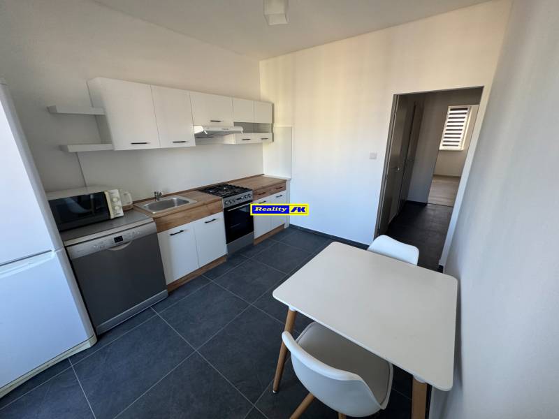 Kuchyně v 3-pokojovém bytě s bílými skříňkami a stolem, na tmavé podlaze.