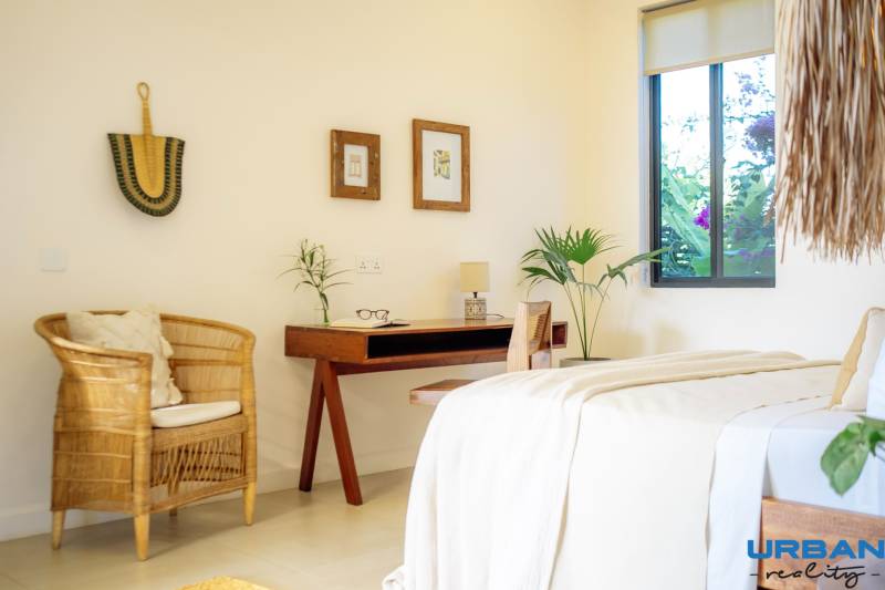 Světlá ložnice v rodinném domě s ratanovým křeslem a dřevěným stolem.