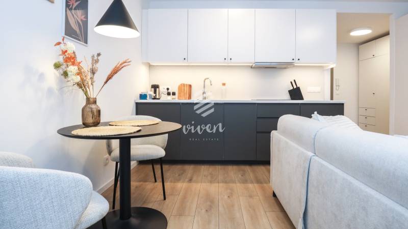 Kuchyně v 1-pokojovém bytě s bílými skříňkami, tmavou linkou a podlahou s dřevěným dekorem.