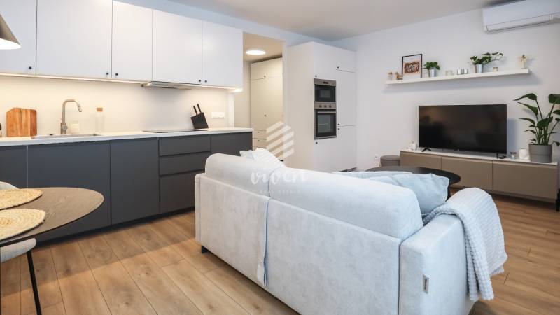 Obývací pokoj jednopokojového bytu s kuchyňskou linkou, gaučem a podlahou s dřevěným dekorem.