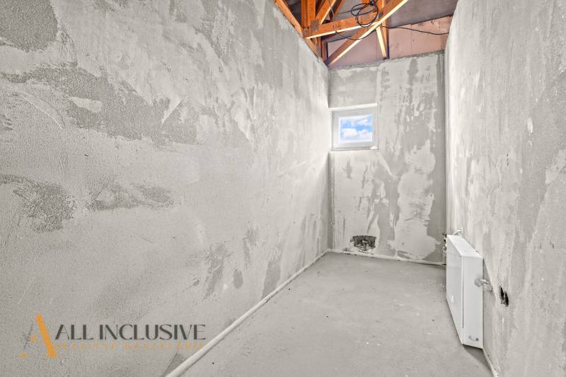 Nedokončená místnost, na stěnách šedá omítka, s malým oknem a viditelnou střešní konstrukcí.