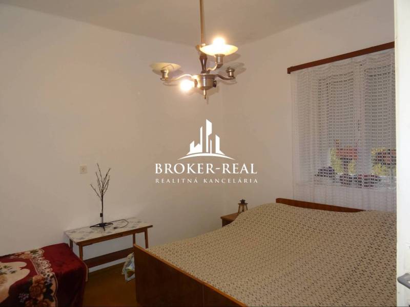 Ložnice v rodinném domě s postelí, stolkem a lustrem, na stěně je okno se záclonou.