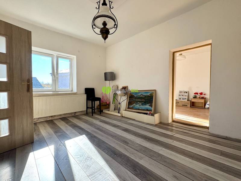 Pokoj v rodinném domě s bílými stěnami, podlahou s dřevěným dekorem a velkým oknem.