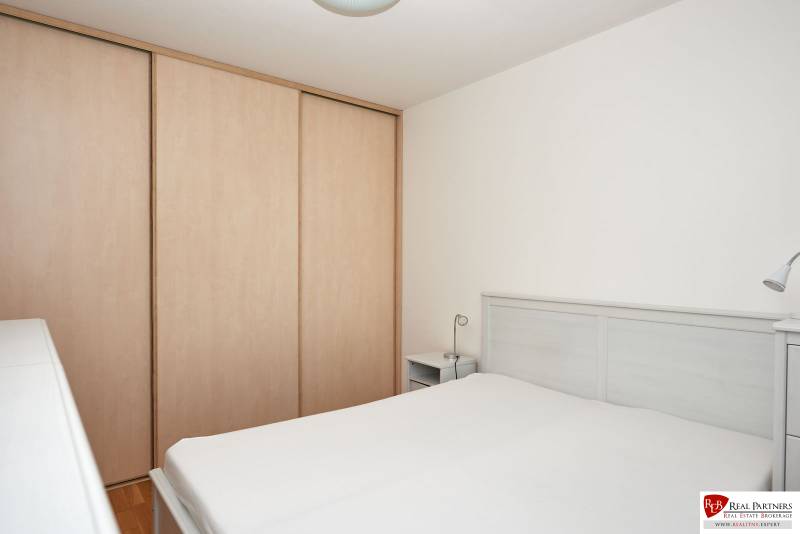 Ložnice v dvoupokojovém bytě s postelí, skříní a bílými stěnami.