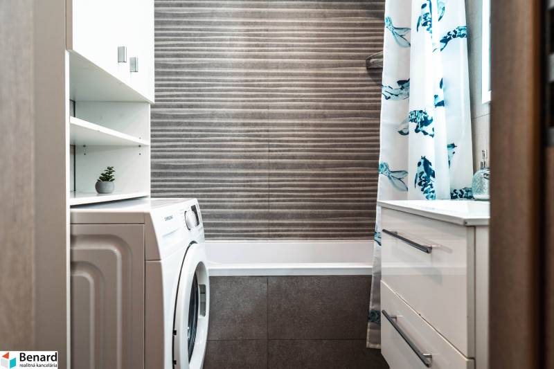 Koupelna v 2pokojovém bytě se sprchovým závěsem, pračkou a úložným prostorem.