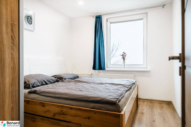 Ložnice s podlahou s dřevěným dekorem a postelí v dvoupokojovém bytě, modrý závěs na okně.