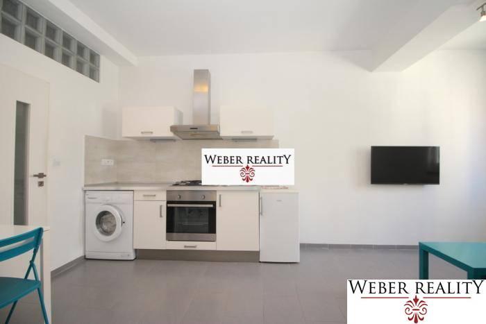 Kuchyně v 2-pokojovém bytě s bílou linkou, pračkou a televizí na stěně.