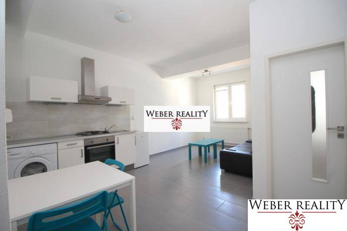 Obývací pokoj v dvoupokojovém bytě s kuchyňským koutem, jednoduchým zařízením a oknem.