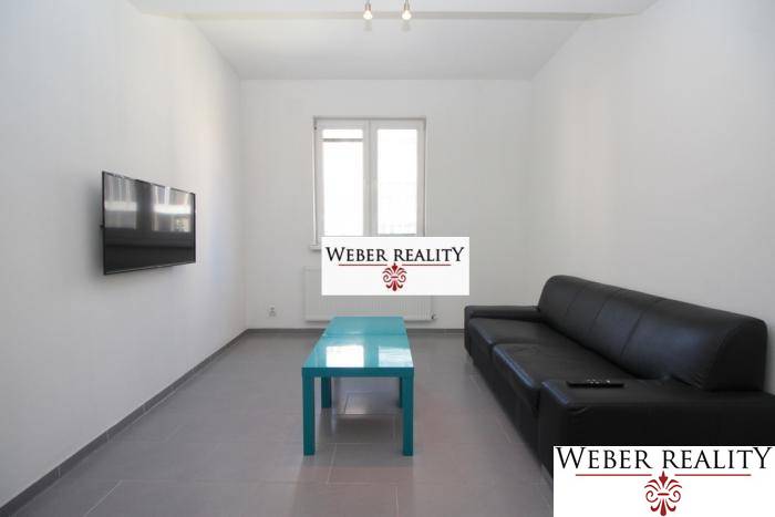 Obývací pokoj dvoupokojového bytu s černou sedačkou, televizorem na stěně a nízkým stolkem.