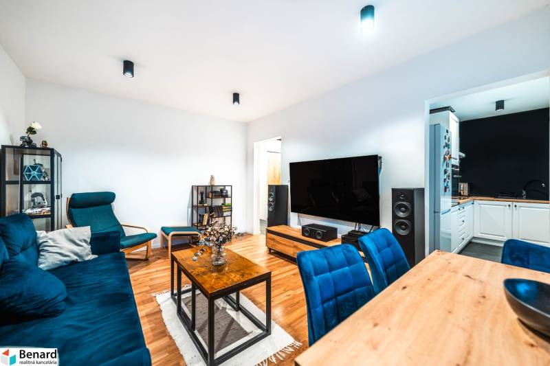 Obývací pokoj dvoupokojového bytu s podlahou s dřevěným dekorem a modrými sedacími soupravami.