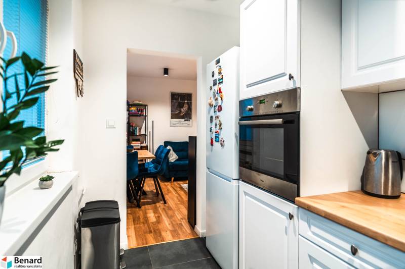 Kuchyňa 2-pokojového bytu s bílými skříňkami, spotřebiči a výhledem do jídelny.