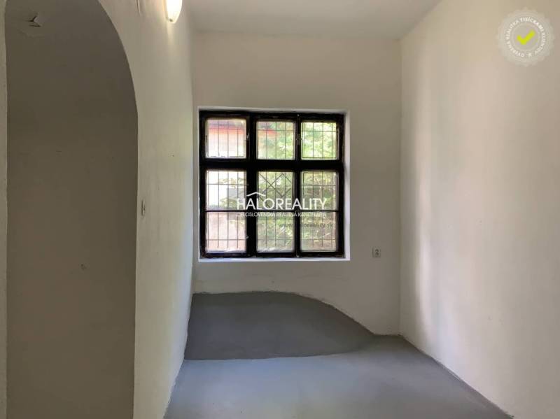 Úzká místnost s bílými stěnami, okno s mříží, šedá podlaha v kanceláři.