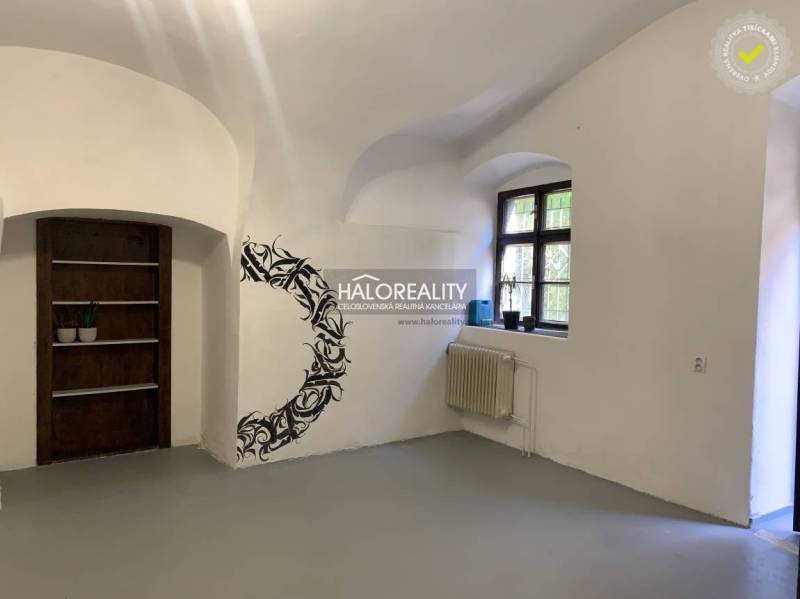 Interiér kanceláře s bílými stěnami, oknem, nástěnnou dekorací a dřevěným regálem.