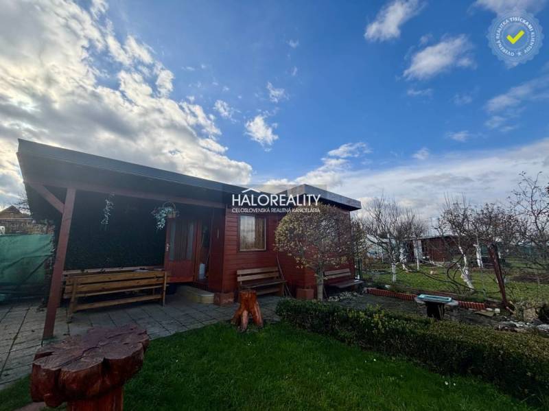 Dřevěná chatka v zahradách Spišské Nové Vsi s lavičkami a květinovým záhonem.