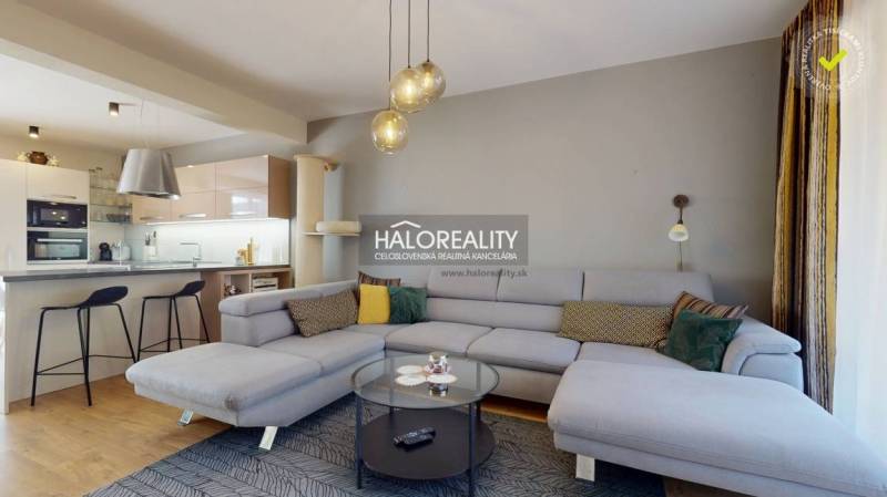 Prostorný obývací pokoj v rodinném domě s pohovkou, kuchyňským koutem a podlahou s dřevěným dekorem.