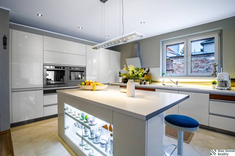 Kuchyně v rodinném domě s centrálním ostrůvkem, bílými skříňkami a spotřebiči.