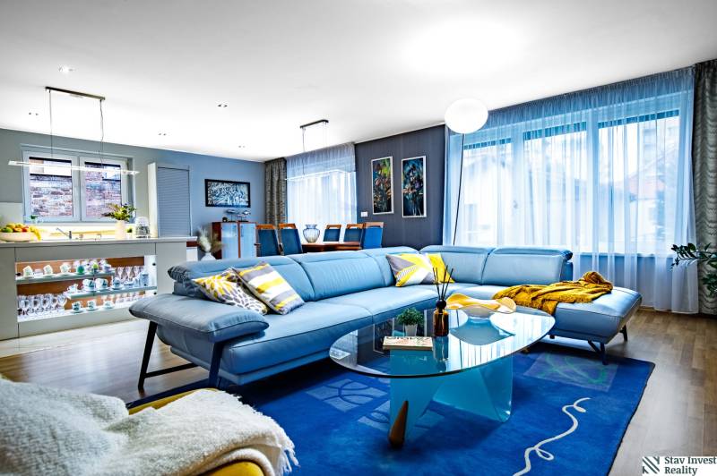 Obývací pokoj v rodinném domě s modrou sedačkou, skleněným stolkem a podlahou s dřevěným dekorem.