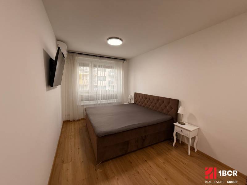 Ložnice s podlahou s dřevěným dekorem, manželskou postelí a televizorem v dvoupokojovém bytě.