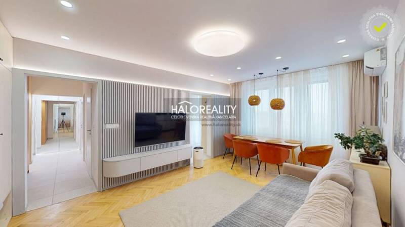 Obývací pokoj v 3-pokojovém bytě s TV na stěně, sedačkou a jídelním stolem.