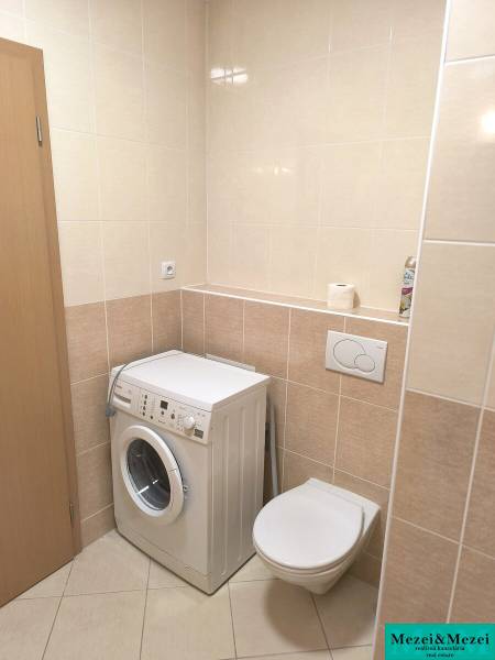 Koupelna v 2-pokojovém bytě se světlými dlaždicemi, pračka a závěsné WC.