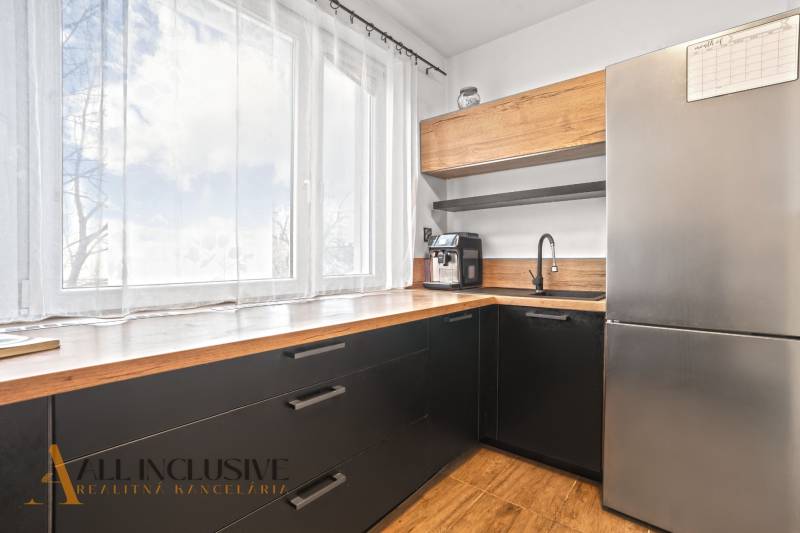 Kuchyně v 3-pokojovém bytě s velkým oknem, černými skříňkami a podlahou s dřevěným dekorem.