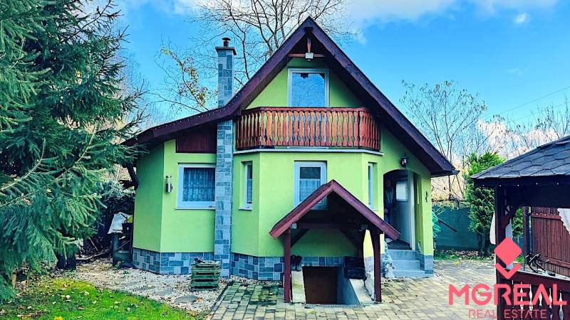 Chata v Partizánském s dřevěným balkónem, obklopená zelení a dlážděným dvorem.